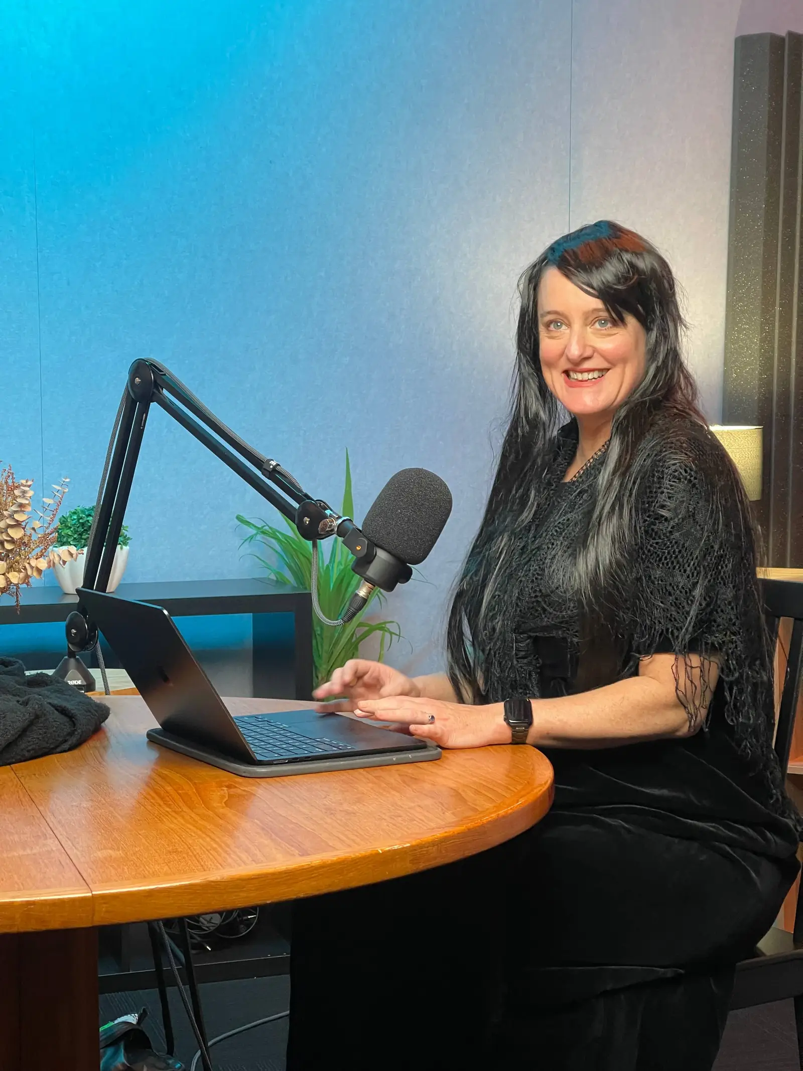 A smiling woman with long dark hair sits at a podcast microphone and laptop at a round table in front of a blue-lit wall.